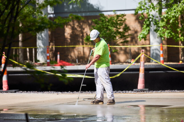 A worker in a hard hat and bright shirt uses a pressure washer to clean a concrete surface, surrounded by caution tape and traffic cones.