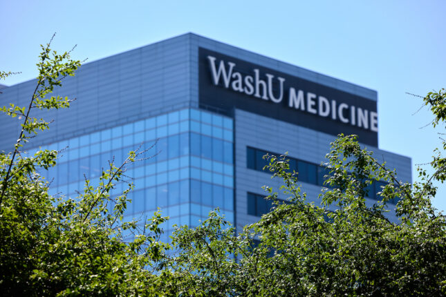 A modern glass building with "WashU MEDICINE" signage is viewed through green tree branches against a clear blue sky.